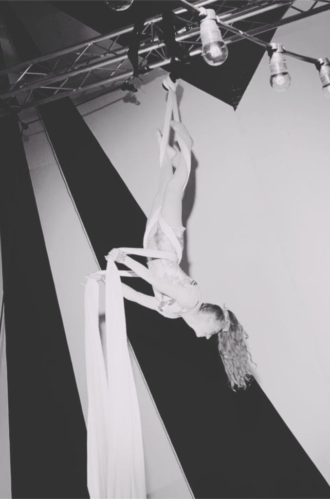 Black and white photo of Freya hanging upside down in white aerial silks.