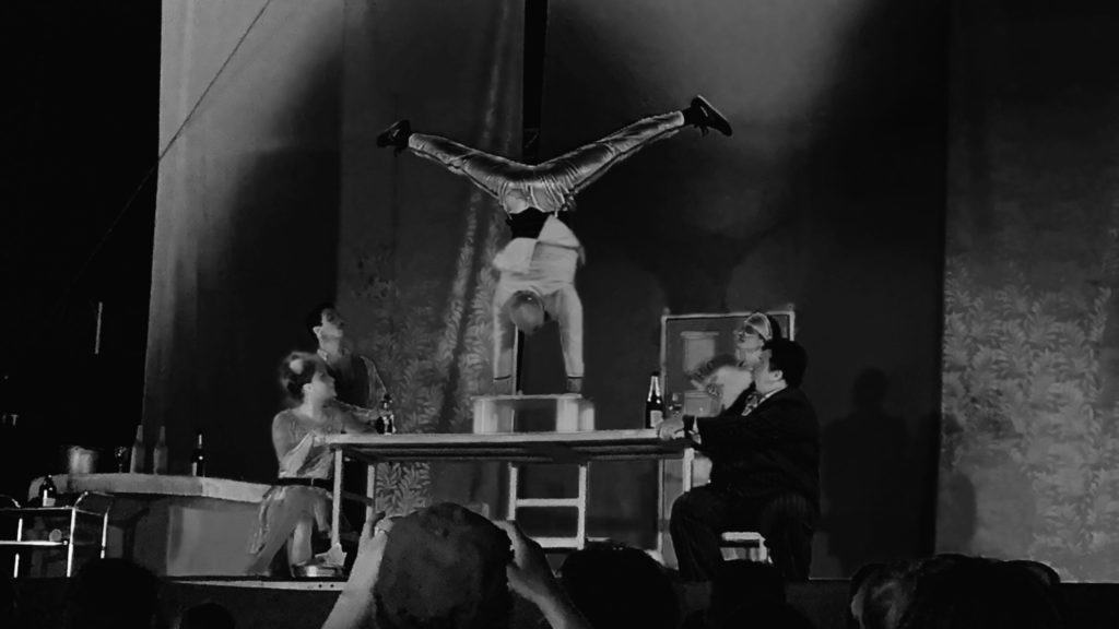 Black and white photo of a man doing a handstand on a table, watched by guests.