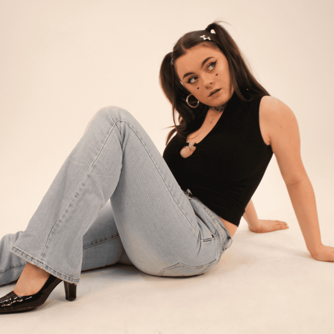 Maisie McGregor, sitting on the floor and looking to the right. She's wearing a black top, grey jeans and black high heeled shoes