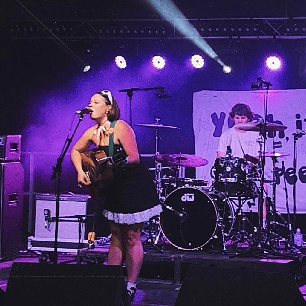 Muffy, January's young creative of the month on a stage singing and playing Guitar. She's joined by a band mate on the drums behind her.