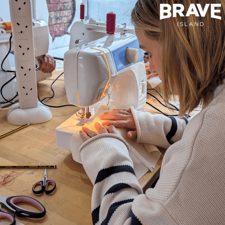 A person with medium length blonde hair sat at a table using a sewing machine