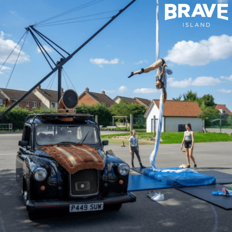 A black "london cab" style car with a metal aerial rig structure coming off the back. From the aerial rig there is an acrobat hanging on silks upside-down whilst two people watch from the ground.