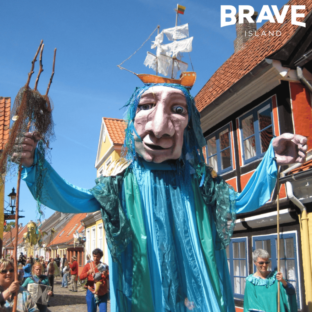 A large Carnival puppet, there's a large head with a model ship on top. The puppet is dressed in blue fabric. It's hands are operated by one person on either side.