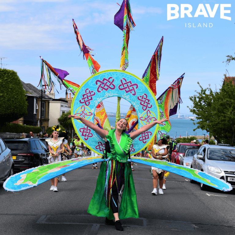 A woman standing in the middle of the road in a large Carnival Costume that extends out to the sides and above her head.