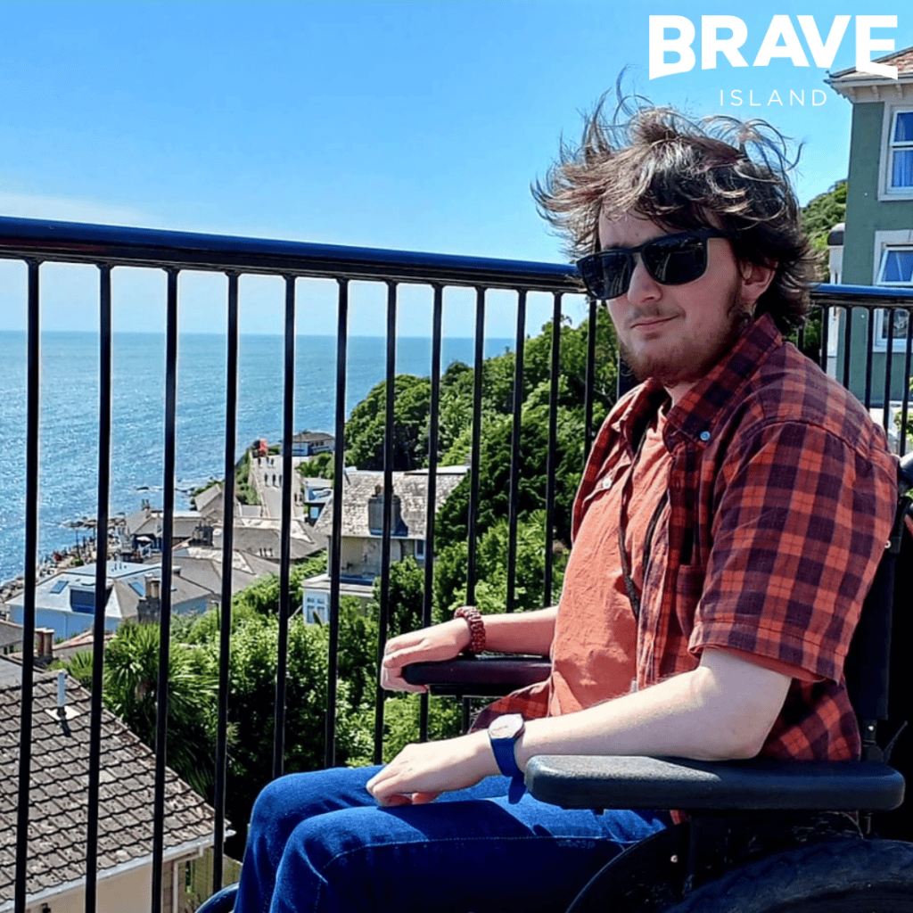 Isaac Leal, a man with dark hair sitting in his wheelchair looking at the camera. In the background is an elevated view of Ventnor Seafront.