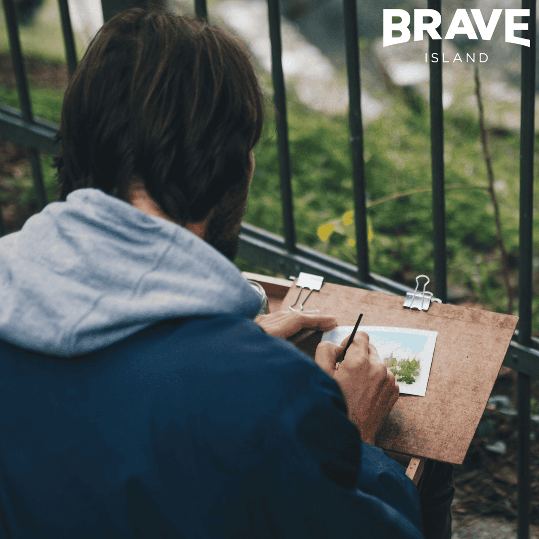 An over the shoulder photo of a man with medium length hair painting a watercolour of a Landscape.