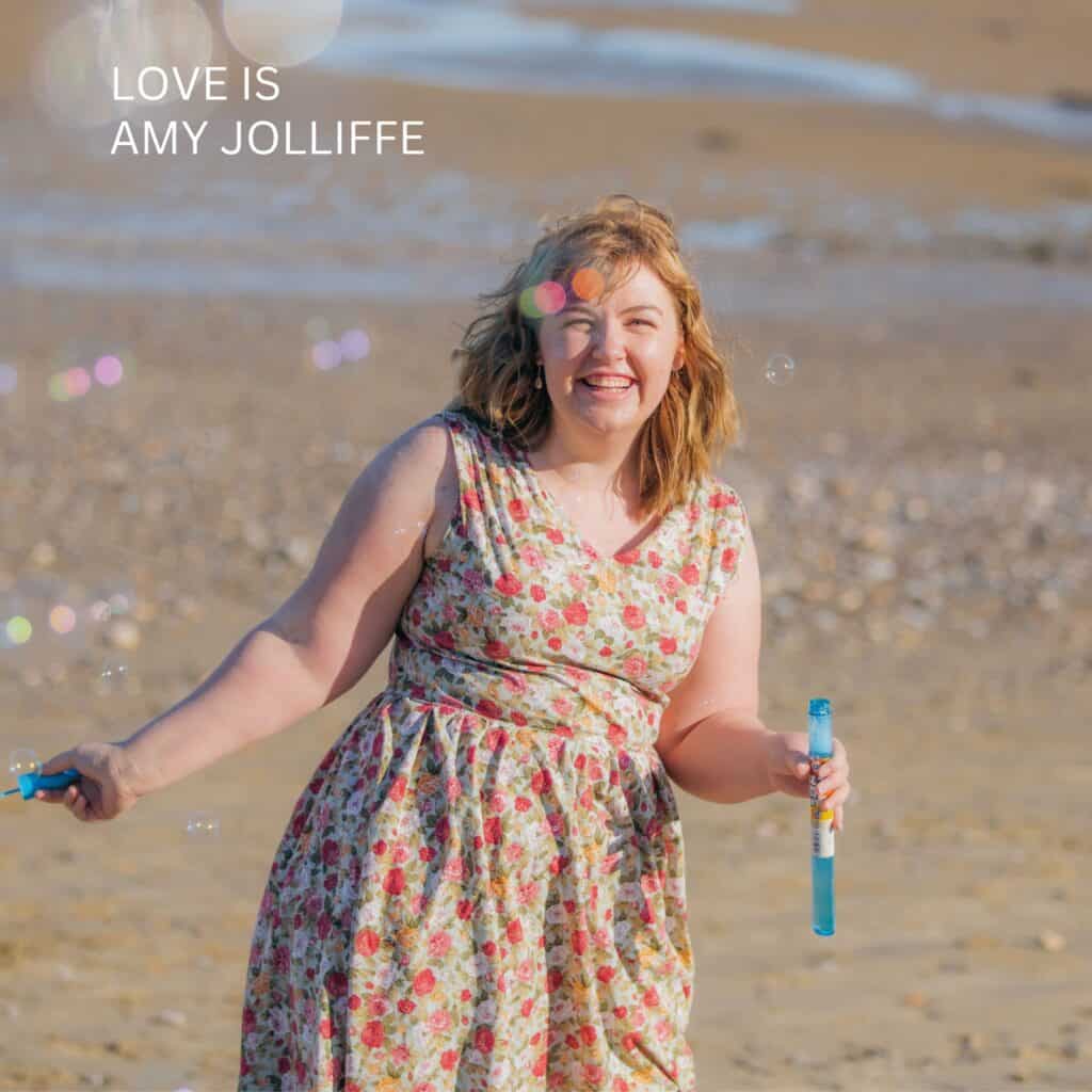 Official Artwork for "Love Is" by Amy Jolliffe. A photo of Amy Jolliffe, on a beach, wearing a floral dress, smiling with bubbles.