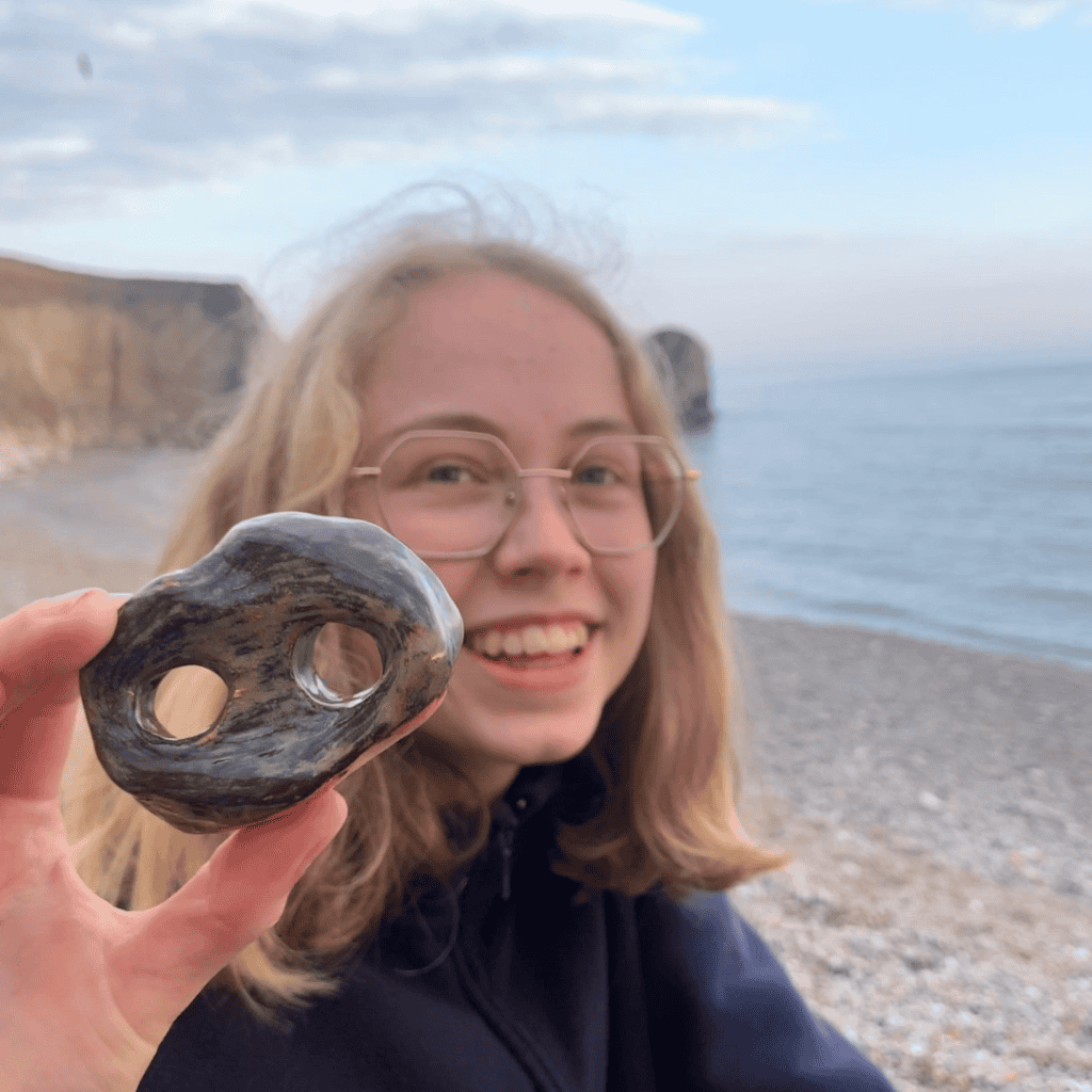 A young girl with blonde hair and glasses on a stony beach holding a stone with two holes up to the camera