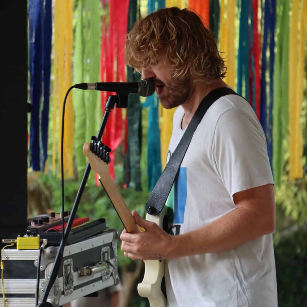 A photograph of a man with medium-length blonde hair and a white tee-shirt singing into a microphone and playing the guitar.
