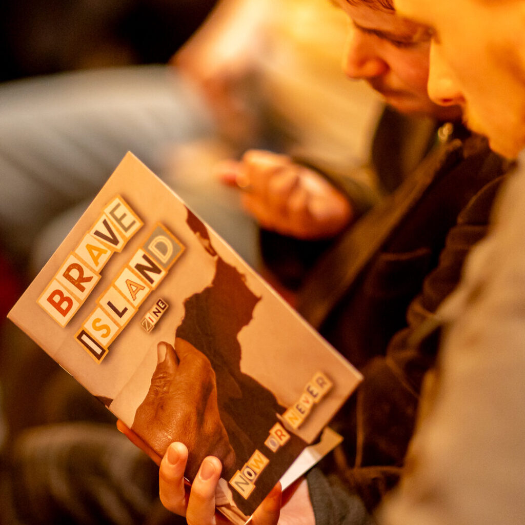 A young man reading the Brave Island Zine, an A5 booklet with multi-coloured cut-out effect lettering spelling "Brave Island Zine Now or Never"