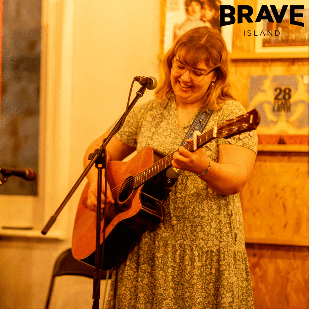 A photograph of Amy Jolliffe, a young woman in a floral dress playing acoustic guitar and singing into a microphone on a stand.