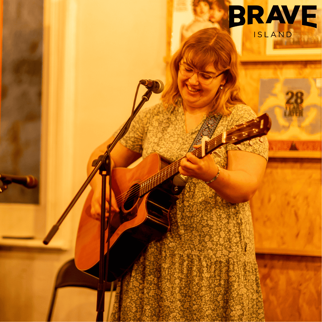 A photograph of Amy Jolliffe, a young woman in a floral dress playing acoustic guitar and singing into a microphone on a stand.
