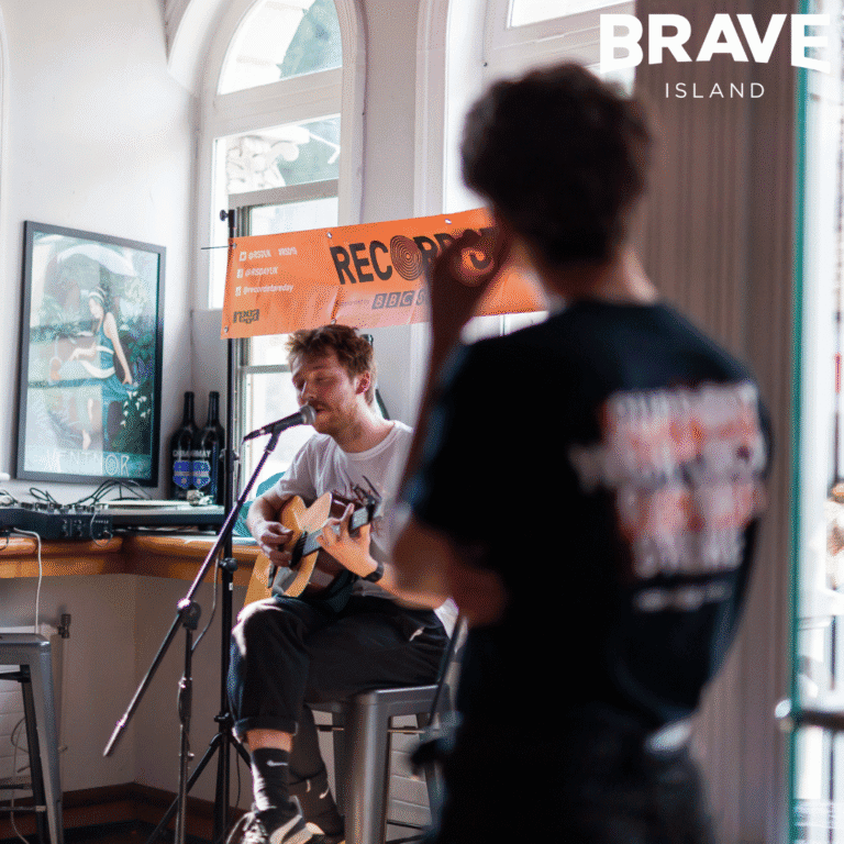 A photograph with a musician playing music on a guitar and singing whilst a young person watches out of focus in the foreground of the image.