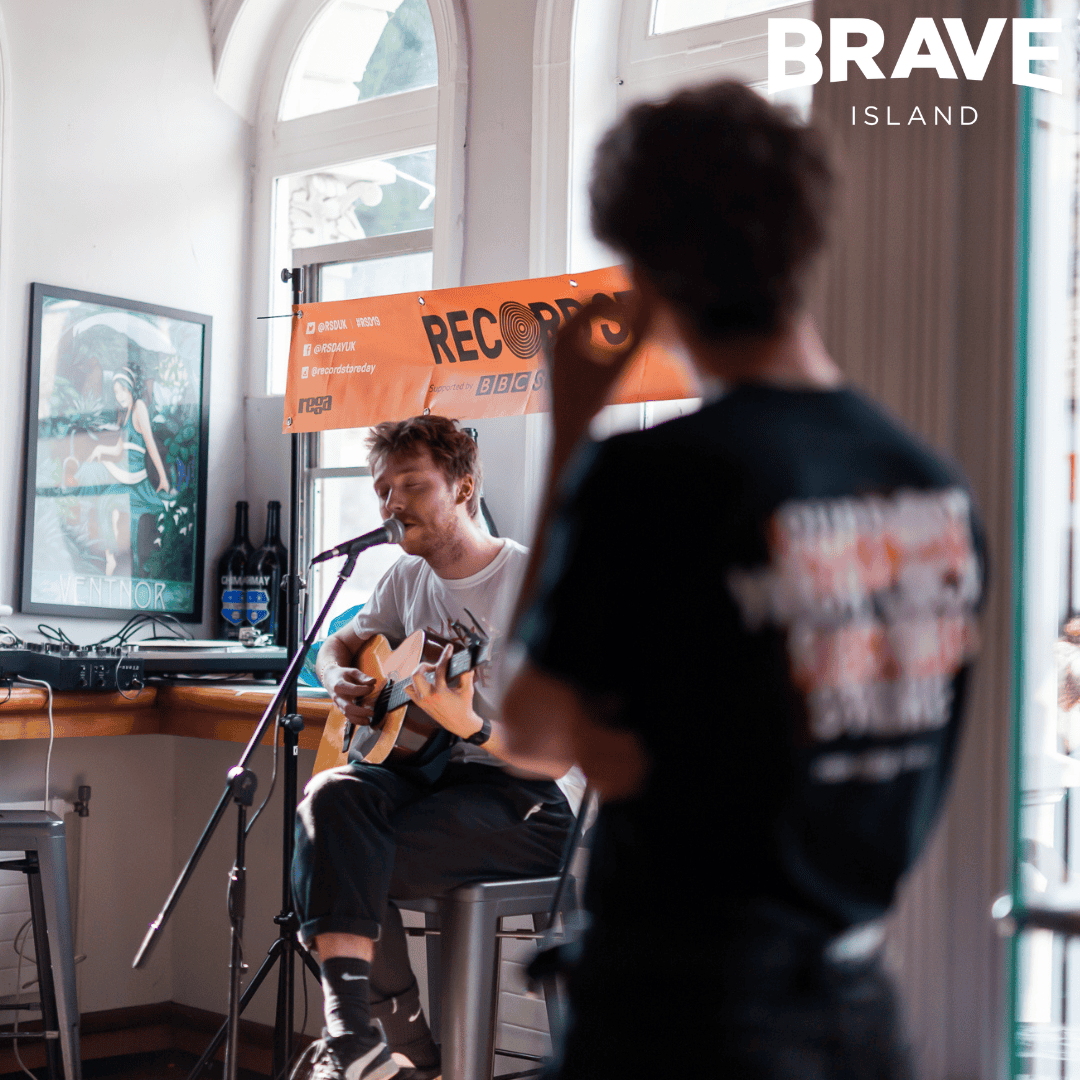 A photograph with a musician playing music on a guitar and singing whilst a young person watches out of focus in the foreground of the image.