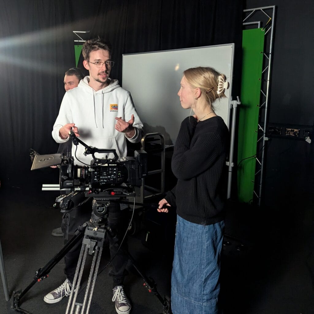 A photograph of two people stood in a TV studio around a large video camera.