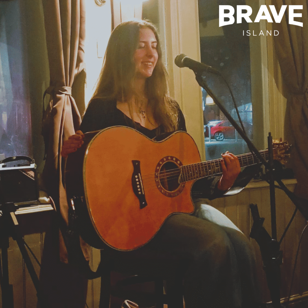 Kyra, a young lady with long dark hair sat on a stool holding a guitar in front of a microphone on a stand.