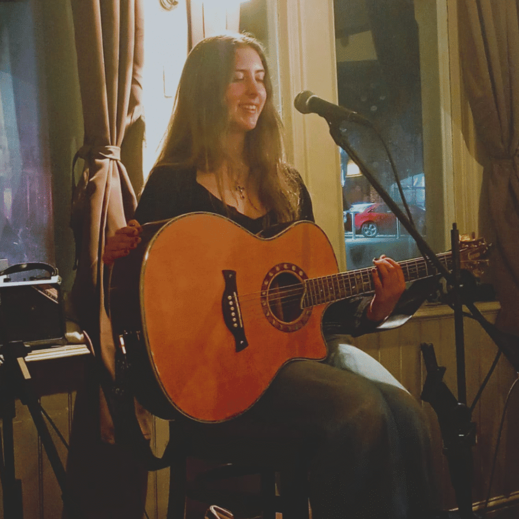 A photo of Kyra, a young lady with long dark hair sat on a stool holding a guitar in front of a microphone on a stand.