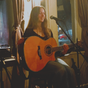 A photo of Kyra, a young lady with long dark hair sat on a stool holding a guitar in front of a microphone on a stand.