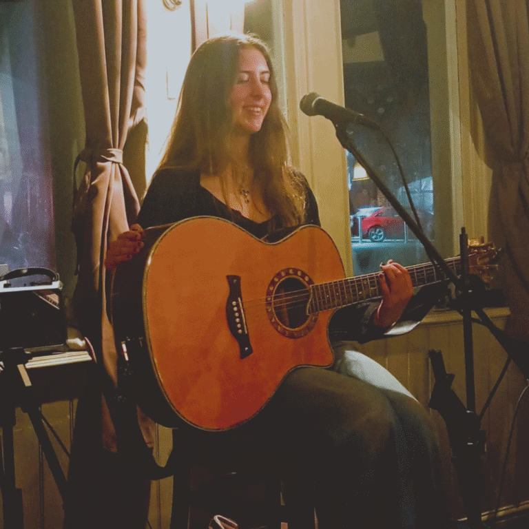 A photo of Kyra, a young lady with long dark hair sat on a stool holding a guitar in front of a microphone on a stand.