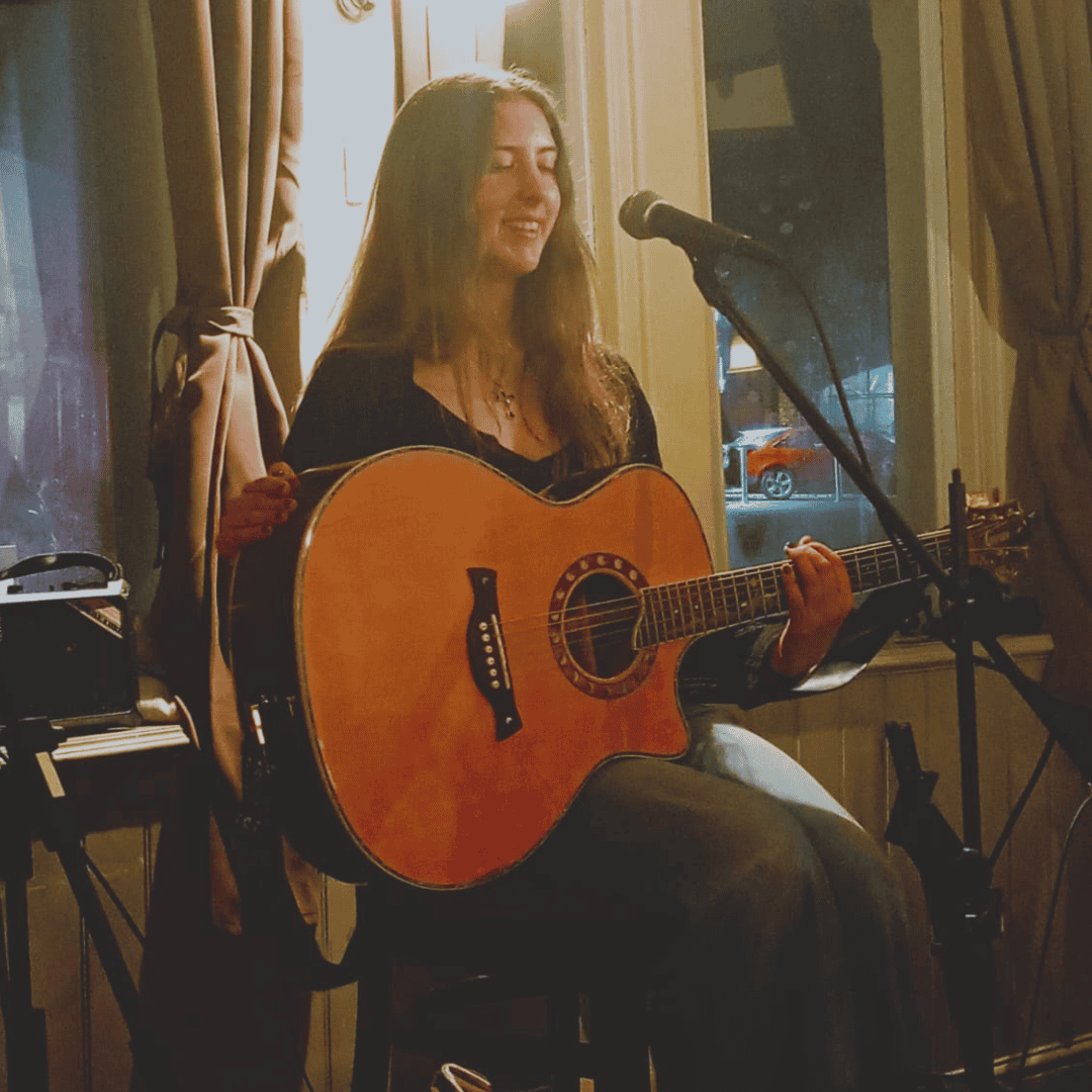 A photo of Kyra, a young lady with long dark hair sat on a stool holding a guitar in front of a microphone on a stand.