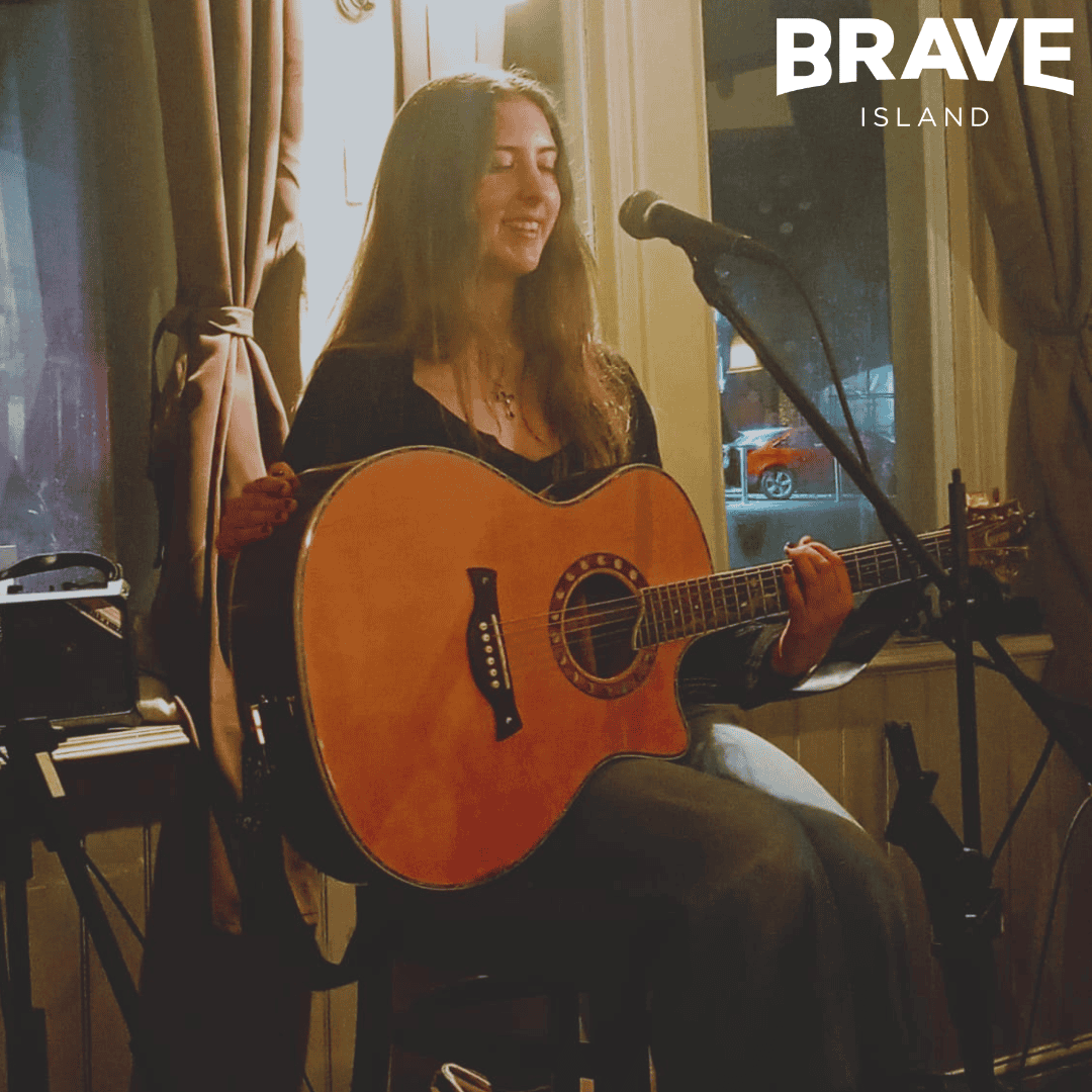 Kyra, a young lady with long dark hair sat on a stool holding a guitar in front of a microphone on a stand.