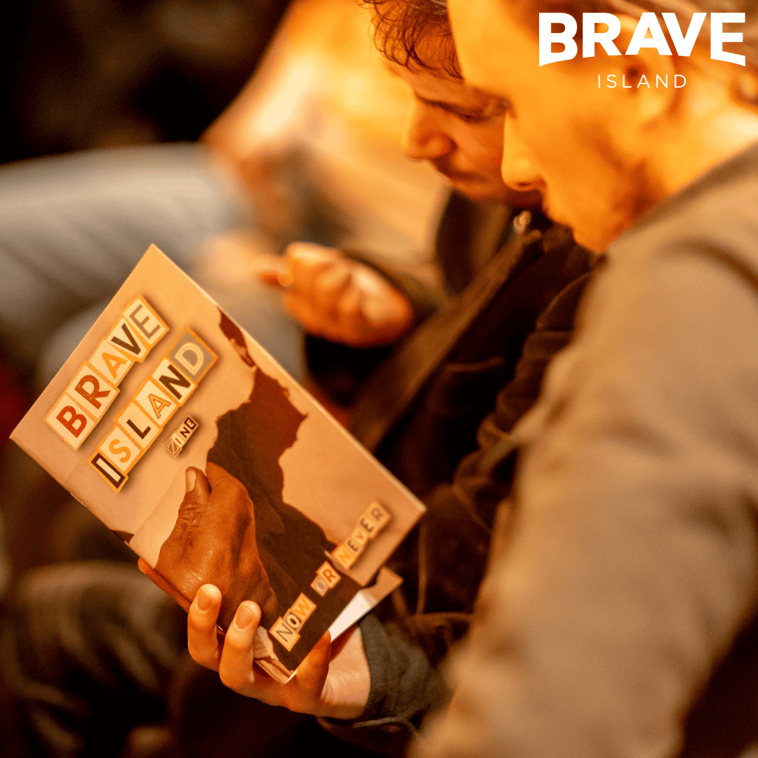 A photograph of a young person, sitting down and reading from a copy of the Brave Island Zine