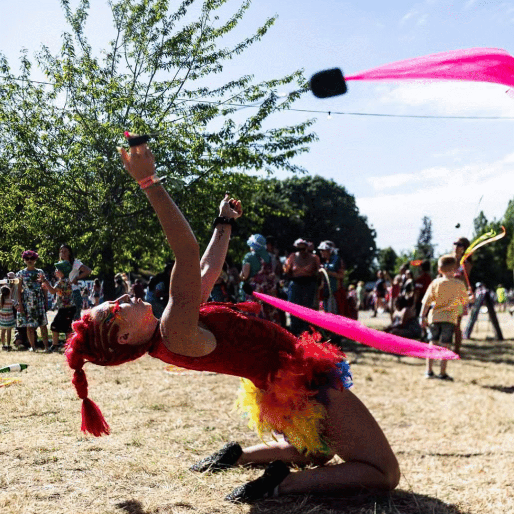 A photograph of a woman spinning Poi. She is dressed in a red bodysuit decorated with multicoloured feathers similar to a parrot. Her hair is shaved at the sides with a red ponytail.