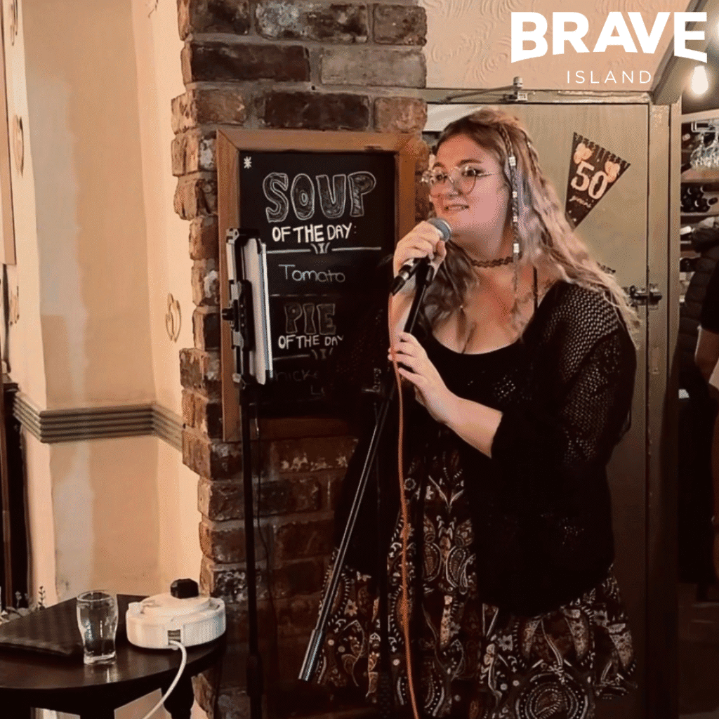 A photograph of Rhi Rhi, a young woman with long blonde hair, singing into a microphone at a pub.
