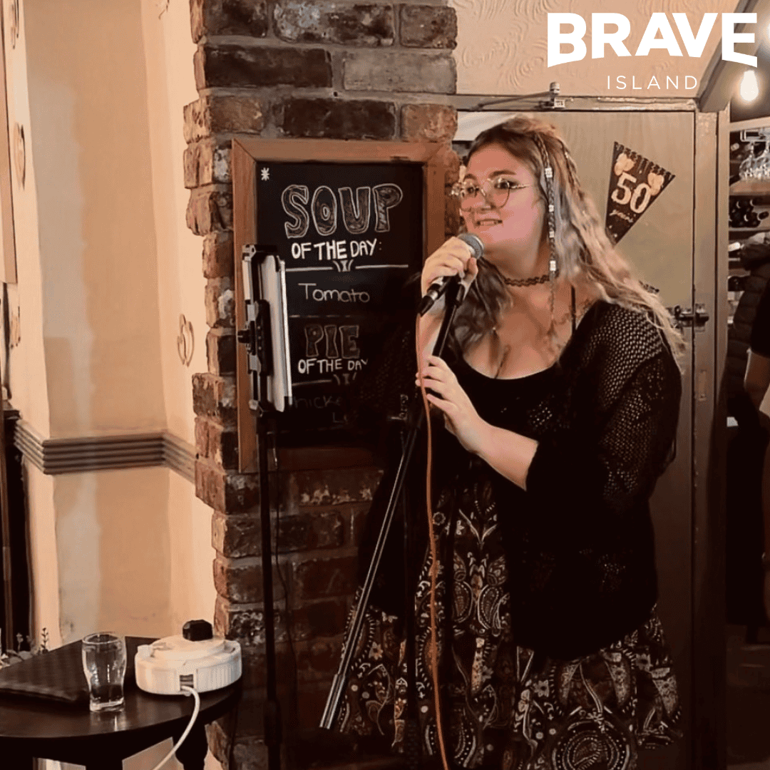 A photograph of Rhi Rhi, a young woman with long blonde hair, singing into a microphone at a pub.
