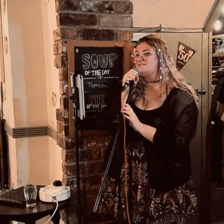 A photograph of Rhi Rhi, a young woman with long blonde hair, singing into a microphone at a pub.