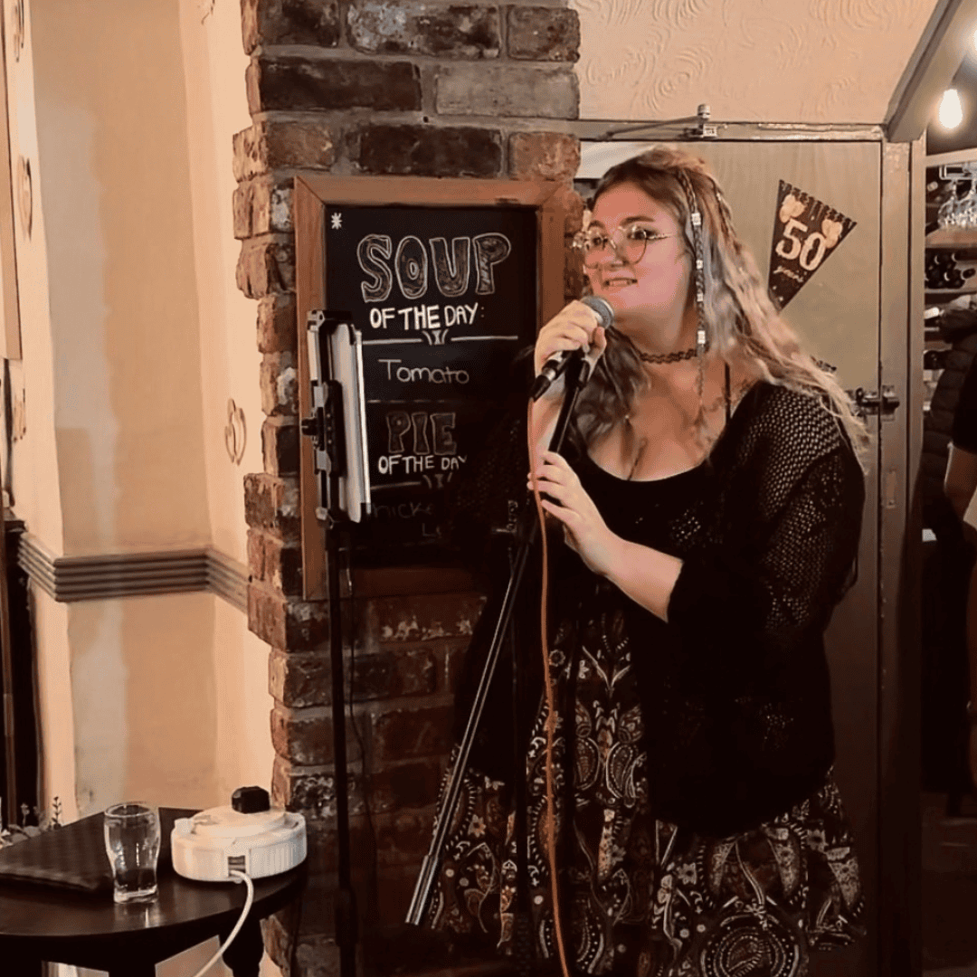 A photograph of Rhi Rhi, a young woman with long blonde hair, singing into a microphone at a pub.